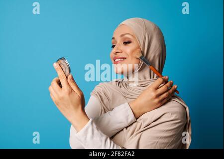 Die schöne arabische muslimische Frau in Hijab hält einen Kosmetikspiegel und einen Make-up-Pinsel und trägt Rouge auf die Wangenknochen ihres Gesichts auf, isoliert auf blauem Ba Stockfoto