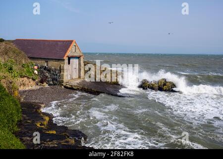 Raues Meer, das bei Flut am Bootshaus der alten Rettungsbootstation an einem windigen Tag auf Felsen stürzt. Moelfre, Isle of Anglesey, Nordwales, Großbritannien Stockfoto