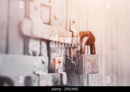 Auf dem grauen alten Holztor befindet sich ein rostiger Riegel, an dem ein altes Metallschloss hängt, das von Sonnenlicht beleuchtet wird. Geschlossen. Stockfoto