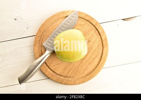 Ein saftiger organischer, reifer, süßer, leuchtend grüner Apfel, auf einem runden Bambustablett mit einem Metallmesser, Nahaufnahme, auf dem Hintergrund eines Tisches aus bemaltem Holz Stockfoto