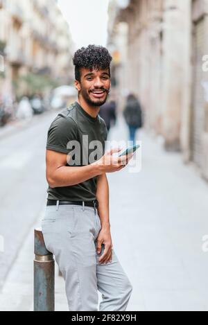 Der junge hispanische Mann lächelt und steht auf der Straße und schaut auf die Kamera. Er trägt grünes T-Shirt und Jeans. Er hält sein Telefon in der Hand. Stockfoto