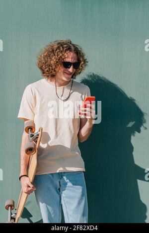 Vertikales Porträt eines langhaarigen jungen Mannes, der sein Telefon überprüft. Er trägt lässige Kleidung, Sonnenbrille und hält ein Skateboard Stockfoto