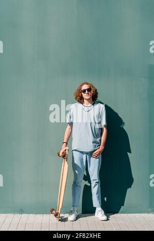 Vertikales Porträt eines kaukasischen jungen Skateboarders, der die Kamera anschaut. Er trägt Freizeitkleidung und Sonnenbrille. Grüner Wandhintergrund Stockfoto
