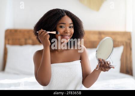 Attraktive afroamerikanische Frau, die im Spiegel schaut und sich zu Hause auf dem Bett schminkt. Tägliches Routinekonzept Stockfoto