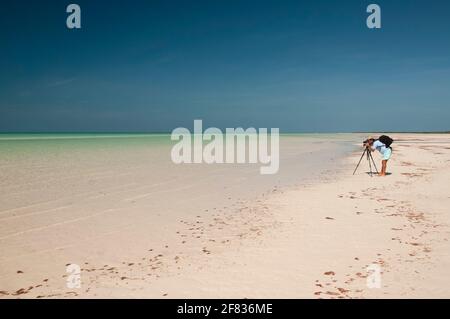 Ein junger Fotograf mit einem Fotostativ an einem einsamen tropischen Strand auf der Insel Holbox in Mexiko bei Ebbe. Im Hintergrund der wolkenlose Stockfoto