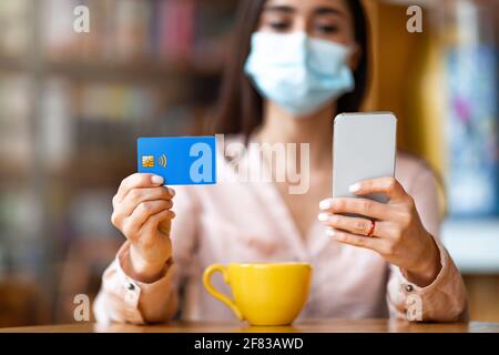 Kontaktlose Bezahlung während der Quarantäne des Coronavirus. Frau in Schutzmaske mit Handy und Kreditkarte Stockfoto