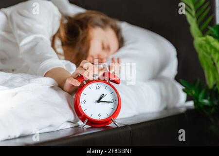 Die Frau, die im Bett schläft, schaltet den Wecker aus, wachen Sie morgens auf, selektiver Fokus. Junge, tausendjährige Hand, die die Schlummertaste auf der roten Uhr im Schlafzimmer drückt Stockfoto