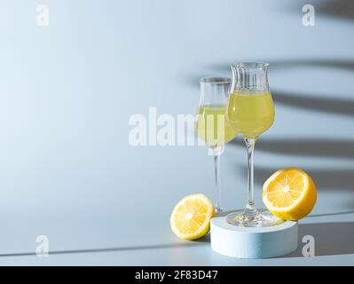 Limoncello, traditioneller italienischer Likör auf hellem Betongrund in den Strahlen der Sonne. Daneben ist eine gelbe Zitrone, frische Zitrusfrüchte. Kompo Stockfoto