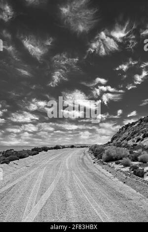 Einsame Wüste Schotterstraße in Richtung Horizont neben Hügeln. Weiße, flauschige Wolken am Himmel. Schwarz und Weiß. Stockfoto