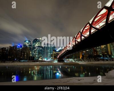 Rote Fußgängerbrücke über den Fluss mit City Skyline bei Nacht und Reflektionen am Snowy Riverbank Stockfoto