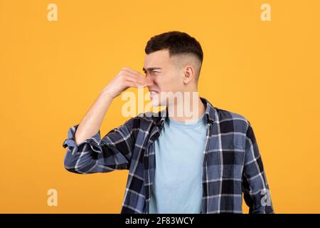 Cooler Tausendjähriger Kerl, der seine Nase schließt, an schrecklichem Geruch leidet und schlechten Geruch auf orangenen Studiohintergrund nicht mag Stockfoto