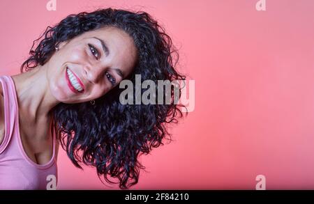 Porträt einer jungen, schwarzen, lockigen Frau, lächelnd, mit ihrem langen Haar zur Seite und Blick auf die Kamera auf einem rosa Hintergrund. Lockiges Mädchen Methode. Haare ca. Stockfoto