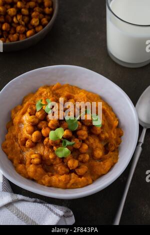 Dicke Kürbissuppe mit gebackenen Kichererbsen und Rettichsprossen auf einem Teller mit einem Glas Milch auf dem Tisch, leckeres gesundes Abendessen Stockfoto