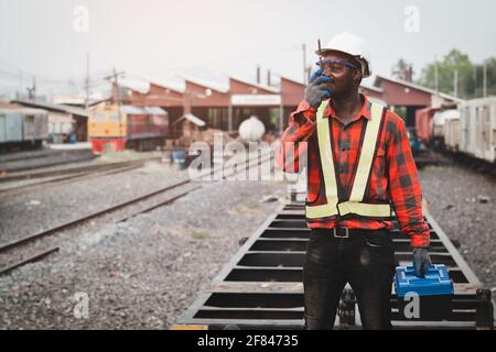 Afrikanischer Maschinenbauingenieur, der mit Walkie Talkie spricht und einen Helm, Haine und Sicherheitsweste trägt, benutzt einen Schraubenschlüssel, um den Zug zu reparieren Stockfoto