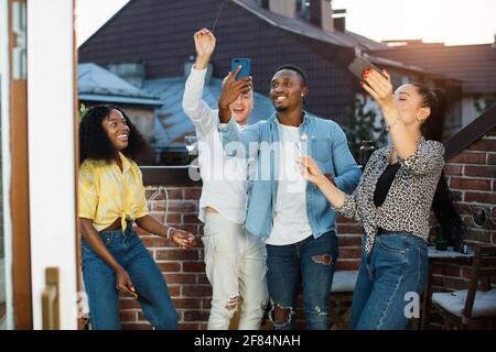 Fröhliche junge Menschen in stilvoller Kleidung mit Smartphones und Wunderkerzen während der Dachparty. Glückliche gemischte Rennfreunde, die ihre Freizeit mit Spaß verbringen. Stockfoto