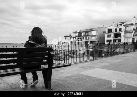 Eine Graustufenaufnahme eines Mädchens, das auf einer Bank sitzt Mit Blick auf Häuser in Calella de Palafrugell Stockfoto