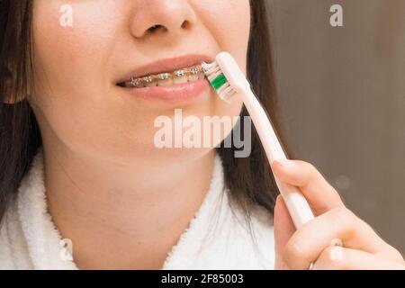 Eine Frau mit Zahnspange oder Halterung putzt sich ihre Zähne mit einer rosa Zahnbürste im Badezimmer. Zahnpflege. Ausrichten und Richten der Zähne. Stockfoto