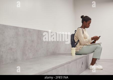 Grafik-Seitenansicht Porträt einer afroamerikanischen Schülerin mit Smartphone, während sie sich in der modernen Schullounge entspannt und Platz für Kopien nutzt Stockfoto