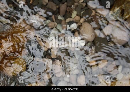 Nahaufnahme des kristallklaren Meerwassers mit Steinen. Hintergrund mit Lichtbrechung auf transparenter Wasseroberfläche mit kleinen Wellen und Wellen, Steine auf Stockfoto