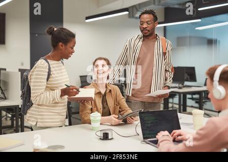 Eine vielfältige Gruppe junger Studenten, die zusammen mit einem Laptop arbeiten und fröhlich chatten, während sie an einem Schulprojekt im College arbeiten, um Platz für Kopien zu erstellen Stockfoto