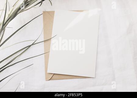 Moderne Sommer Schreibwaren Stillleben. Trockenes Palmblatt auf weißem Leinentischtuch. Leere Grußkarte imitieren Szene mit Handwerk Umschlag in Sonnenlicht. Flach Stockfoto