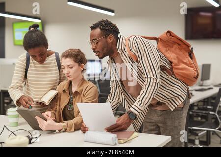 Eine vielfältige Gruppe von Schülern, die in einer modernen Schulbibliothek zusammenarbeiten Und mit einem digitalen Tablet Stockfoto