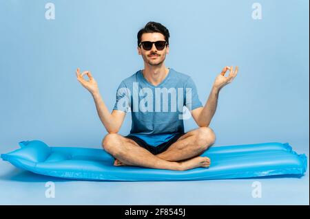 Charismatischer kaukasischer zufriedener Kerl mit Sonnenbrille, barfuß, sitzt auf einem aufblasbaren blauen Kreis in Lotusposition, meditiert, träumt von einem Urlaub und vom Meer. Konzept der Ruhe und Harmonie Stockfoto