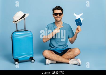 Kaukasischer fröhlicher männlicher Tourist in Sonnenbrillen und Sommerkleidung sitzt auf isoliertem blauem Hintergrund, hält Reisepass und kleines Flugzeug in der Hand und zeigt Finger darauf, blauer Koffer steht in der Nähe und lächelt Stockfoto