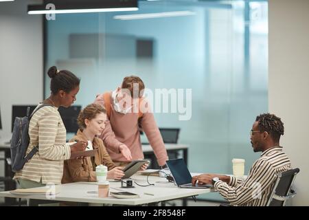 Seitenansicht auf verschiedene Gruppe von Studenten, die in der modernen Schulbibliothek zusammenarbeiten und Computer verwenden, Kopierraum Stockfoto