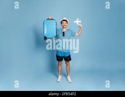 Ein Foto in voller Länge von einem fröhlichen, aufgeregt kaukasischen Kerl in einem Hut hält einen blauen Koffer und ein kleines Flugzeug, männlicher Tourist schaut direkt auf die Kamera, lächelnd glücklich, stehen auf einem isolierten blauen Hintergrund Stockfoto