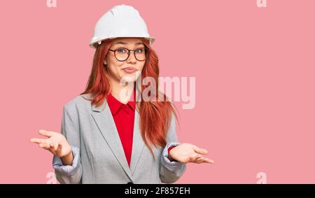 Junge Rotschopf Frau trägt Architekt Hardhat ahnungslos und verwirrt Ausdruck mit Armen und Händen angehoben. Zweifel Konzept. Stockfoto