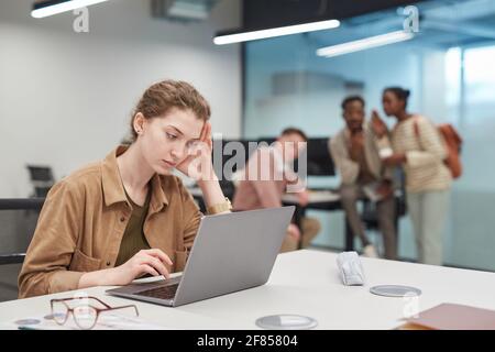 Porträt einer gestressten jungen Frau, die im Büro einen Laptop benutzt oder einen Coworking Space mit einer Gruppe von Menschen im Hintergrund, einem Kopierraum Stockfoto