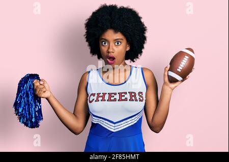 Die junge afroamerikanische Frau, die in einer Cheerleader-Uniform Pompon und Fußballball im Schockgesicht hielt und skeptisch und sarkastisch aussahen, überraschte wi Stockfoto