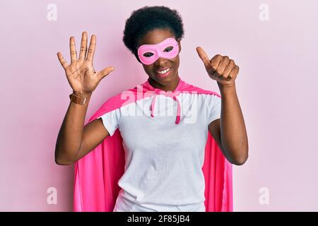 Junge afroamerikanische Mädchen trägt Superhelden-Maske und Cape-Kostüm zeigt und zeigt mit den Fingern Nummer sechs, während sie selbstbewusst und glücklich lächelt. Stockfoto