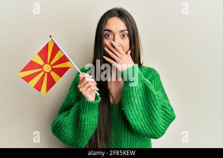 Junge hispanische Mädchen hält mazedonische Flagge über den Mund mit der Hand, schockiert und Angst vor Fehler. Überrascht Ausdruck Stockfoto