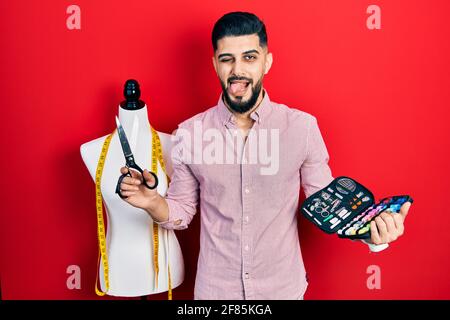 Schöner Schneider Mann mit Bart hält Näh-Kit und Schere ragt Zunge heraus glücklich mit lustigen Ausdruck. Stockfoto