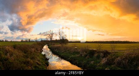 Ein lebhafter und dramatischer Sonnenuntergang über dem Lake Ditch nach einem Sturm Anfang April. Der Himmel spiegelt sich im Wasser. Der Graben ist eine führende Linie. Stockfoto