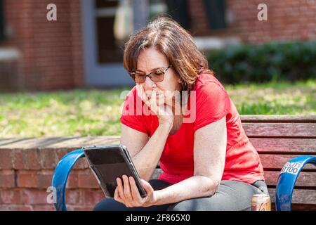 Frederick, MD, USA 04-07-2021: Nahaufnahme Isoliertes Bild einer älteren kaukasischen Frau, die ein Buch oder eine Zeitung von ihrem eBook Reader Tablet auf einem sonnigen Platz liest Stockfoto