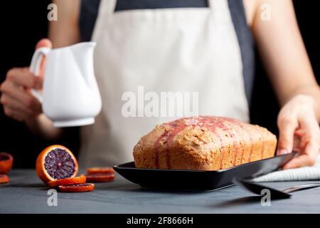 Nahaufnahme Bild von frischen hausgemachten Blut-Orange-Kuchen in einer Laib-Pfanne gemacht und serviert auf schwarzem Porzellan-Teller auf dunklem Stein Hintergrund. Eine Frau gießt Bloo aus Stockfoto