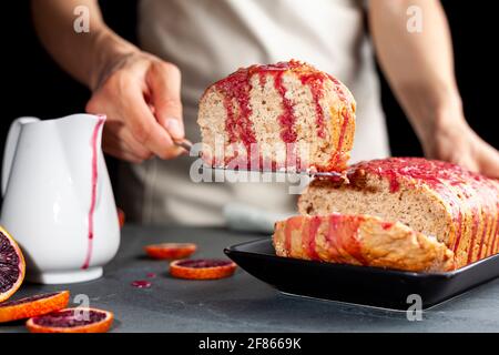 Nahaufnahme Bild von frischen hausgemachten Blut-Orange-Kuchen in einer Laib-Pfanne gemacht und serviert auf schwarzem Porzellan-Teller auf dunklem Stein Hintergrund. Eine Frau dient Stockfoto