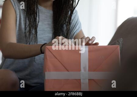 Frau mit langen Haaren in Zöpfen, die einen Bogen binden Eine Geschenkbox in Nahaufnahme an ihren Händen In einem schwach beleuchteten Raum Stockfoto