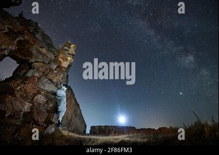 Spaceman in weißem Anzug auf Ruine sitzend und Blick auf schönen Sternenhimmel in der Nacht. Konzept des kosmischen Reisenden. Stockfoto