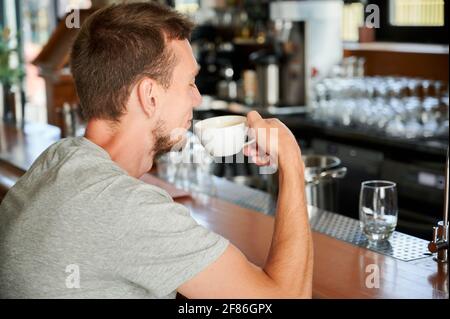 Rückansicht eines hübschen jungen Mannes, der an der Bar sitzt und im Café eine Tasse Kaffee trinkt. Konzept der Ruhe mit heißem Getränk in guter Laune. Stockfoto