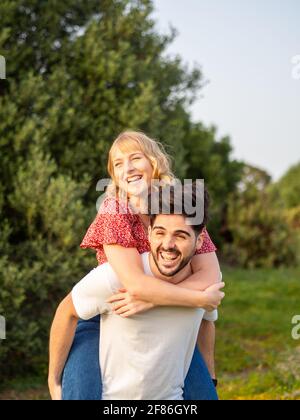 Das junge spanische Paar hat Spaß im Park; ein Kerl gibt seiner Freundin eine Huckepack-Fahrt Stockfoto