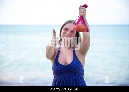 Frau hält Mesh-Tasche mit Obst und zeigt Daumen nach oben. Auf dem Hintergrund ist Meer oder Meer. Stockfoto