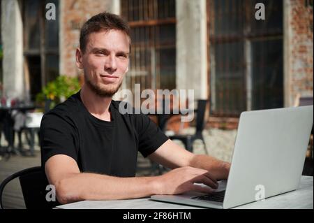 Porträt eines lächelnden jungen Studenten, der an der frischen Luft auf der Terrasse in einem Laptop arbeitet oder studiert. Konzept der Zeit auf der Straße im Café mit Computer. Stockfoto
