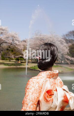 Junge Frau im orangefarbenen Kimono, die den Brunnen im sonnigen Frühling beobachtet parken Stockfoto