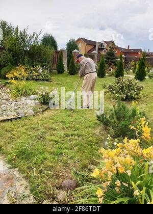 Ein älterer Mann von 80 Jahren arbeitet im Garten. Der Gärtner reckt das gemähte Gras mit einem Rechen. Aktives Alter. Arbeiten im Garten in Quarantäne in der c Stockfoto