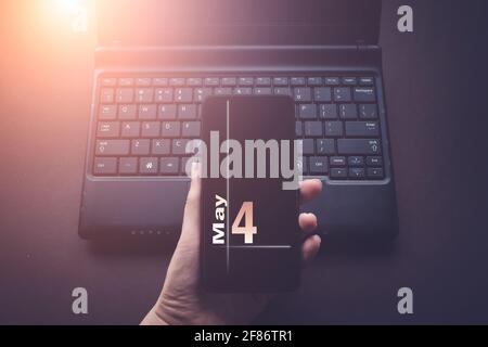 Mai. Tag 4 des Monats, Kalenderdatum. Hand halten Handy auf Laptop-Computer dunklen Hintergrund mit Sonnenschein. Frühling Monat, Tag des Jahres Konz Stockfoto