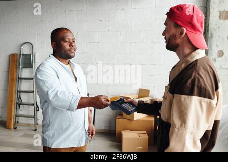 Junger Lader in Arbeitskleidung hält Terminal während afrikanische männliche Client Zahlung für das Laden und Bereitstellen des Services per Kreditkarte Stockfoto